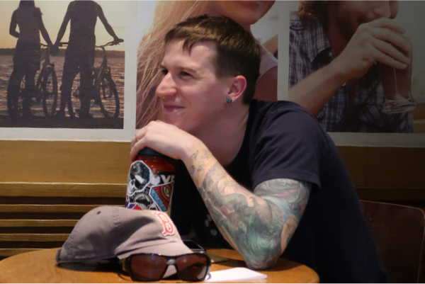 Young man with tatoos sitting at a coffee shop leaning on a water bottle smiling