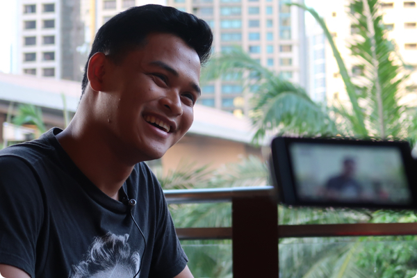 A young man smiling with a backdrop of tropical plants and city buildings. An out of focus camera records the interview in the foreground.