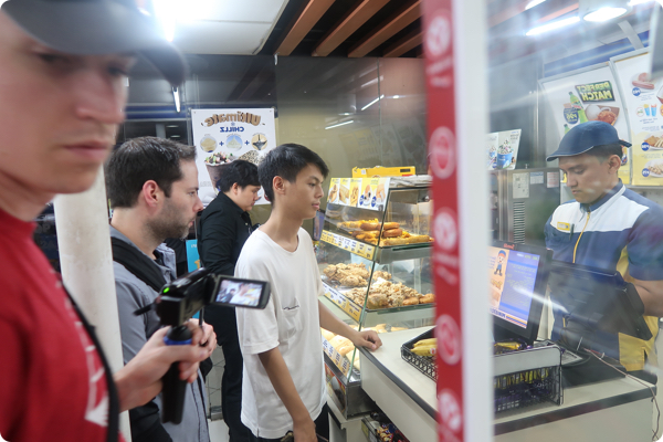 A young college student waits in line in a busy conveinence store. The store clerk processes his transaction for prepaid data. A young man stand near the camera recording the interaction.